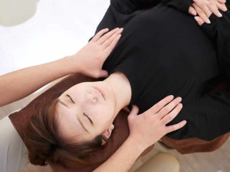 A person lies on a brown towel with eyes closed while another gently places their hands on the person's chest and shoulder, possibly performing a Neck & Shoulders Massage or other therapeutic treatment focused on relaxation.