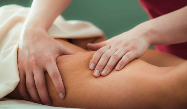 A close-up of a person receiving a Head, Neck & Shoulders Massage. A pair of hands gently presses and massages the person's bare back, with a neutral-colored sheet partially covering them. The background is softly blurred.